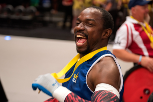 Ein Mann im Rollstuhl mit ausgestreckten Armen und einer Medaille um den Hals lächlt im Vordergrund, im Hintergrund sind Menschen, Stühle und andere Gegenstände zu sehen.