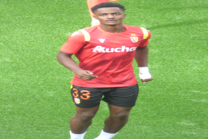 Emmanuel Adebayor, ein Mann in einem roten T-Shirt und schwarzen Shorts, geht auf einem Füßballfeld mit zwei Personen im Hintergrund.