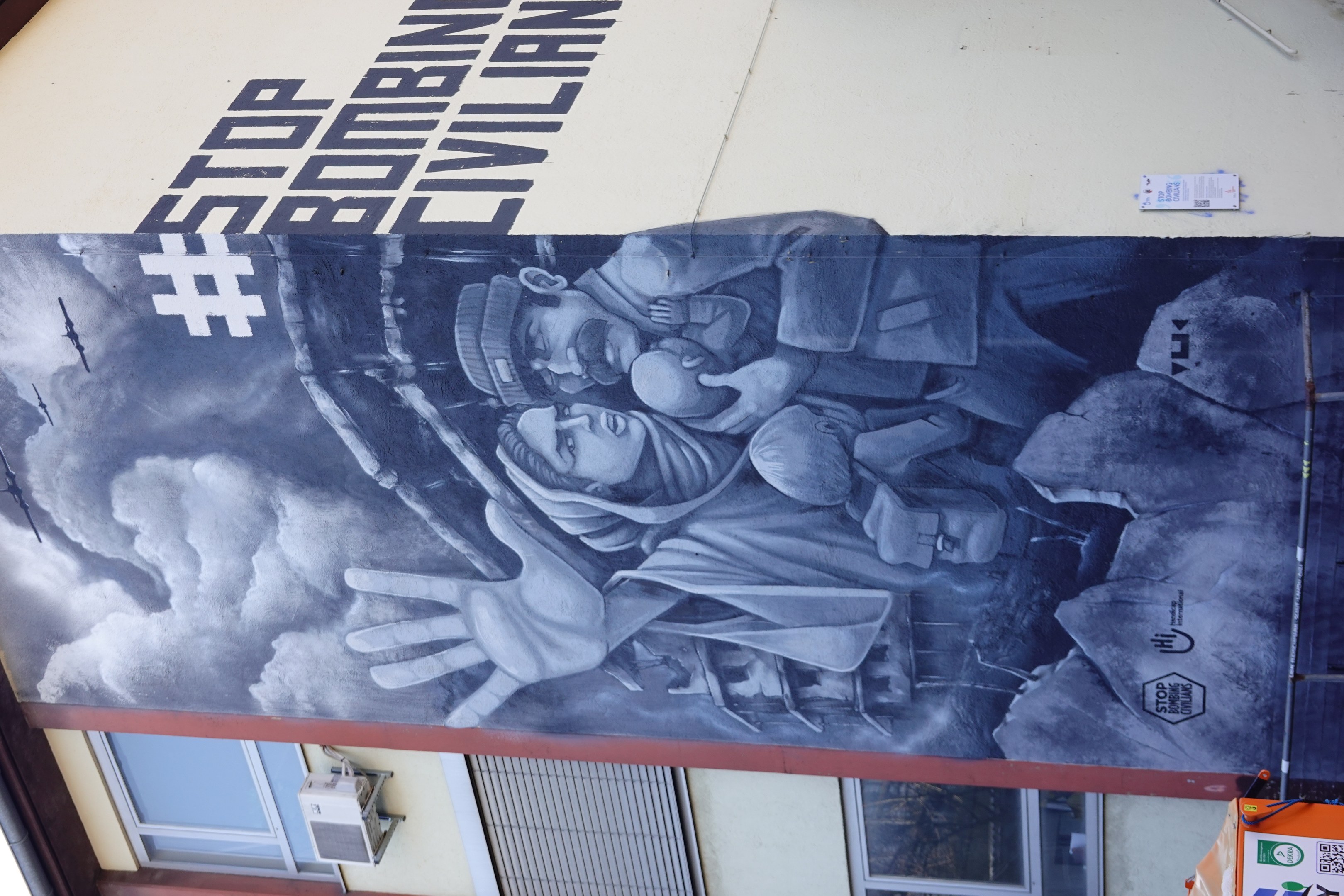 Mural an einer Gebäudeseite mit der Aufschrift "Stoppt das Bomben von Zivilisten" und einem gemalten menschlichen Gesicht über Glasfenster und andere Gegenstände.