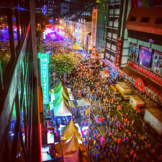 Ein lebhaftes Karnevalsszenario mit einer großen Menschenmenge auf der Straße, umgeben von Zelten, Geschäften, Gebäuden und Bannern, mit einer hell erleuchteten Fläche im Hintergrund.