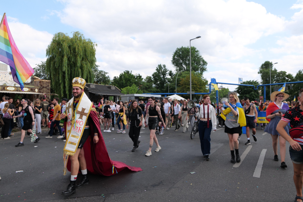 Eine Gruppe von Menschen bei der 2018er Gay Pride Parade mit einer Regenbogenflagge und Musikinstrumenten, einige tragen Mützen, vor dem Hintergrund von Laternenmasten, Bäumen, Schuppen und einem bewölkten Himmel.