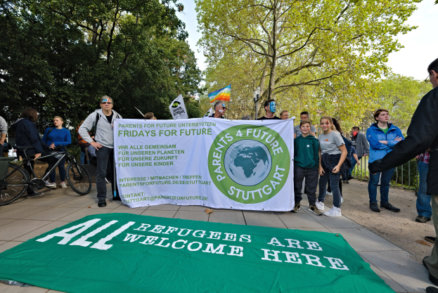 Eine Gruppe von Menschen, die auf dem Boden stehen, einige halten ein Banner mit der Aufschrift "Alle Flüchtlinge sind hier willkommen" und eine Flagge, mit Fahrrädern, einem Zaun, einem Straßenpfosten, einer Schautafel, Bäumen und einem bewölkten Himmel im Hintergrund.