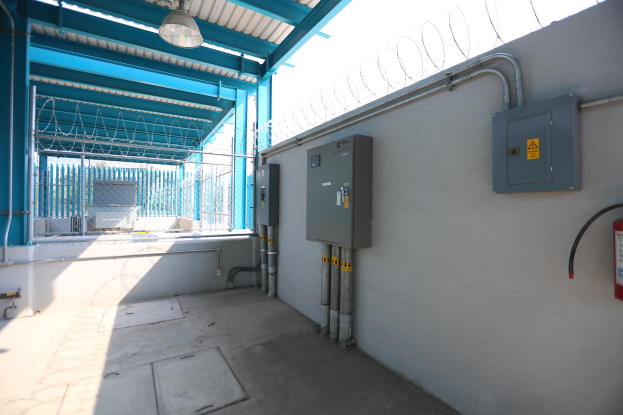 Ein Raum in einer elektrischen Umspannstation mit einem Feuerlöscher auf der rechten Seite, elektrischen Kästen und Rohren an der Wand, einer Decke mit einer Lampe oben und einem Zaun im Hintergrund.