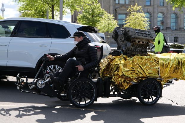 Ein Mann im Rollstuhl mit einem großen Motor auf dem Rücken, umgeben von Fahrzeugen auf einer Straße mit Bäumen, Gebäuden, Polen und einem klaren blauen Himmel im Hintergrund, trägt eine schwarze Jacke und eine Kappe und hält ein Objekt.