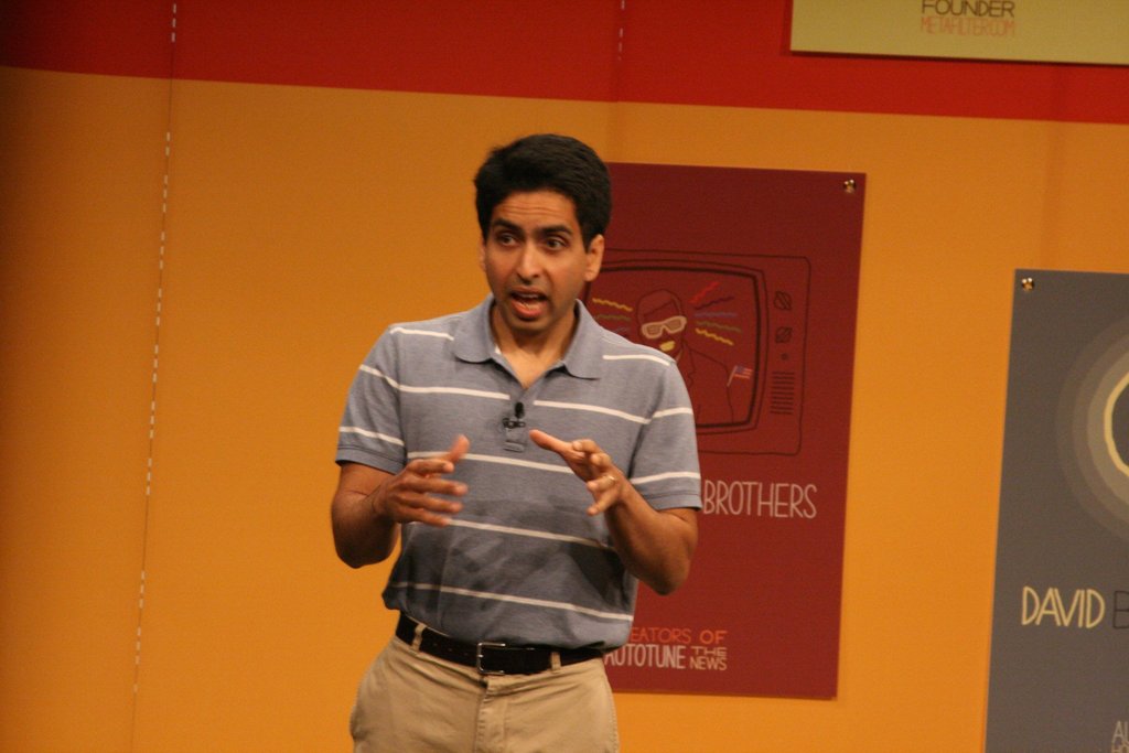 Ein Mann steht auf einer Bühne und spricht vor einer Wand mit Plakaten.