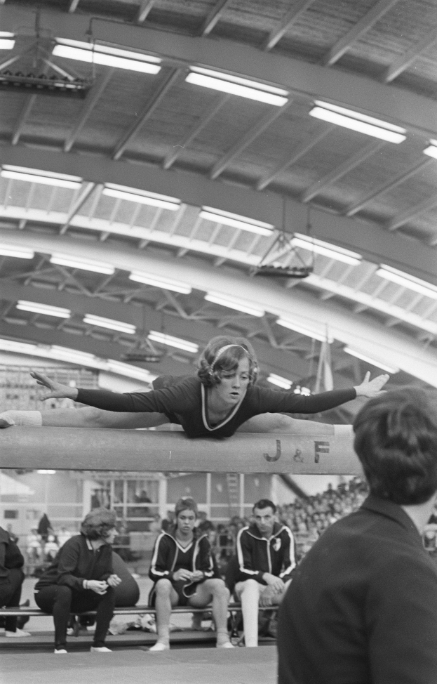 Eine Frau führt Gymnastik auf einem Balancebalken in einer Turnhalle aus, umgeben von Zuschauern auf Bänken und einigen Stehenden unter Deckenlampen, dargestellt in Schwarz-Weiß.