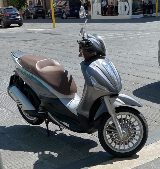 Ein Roller mit einem Helm darauf auf einer Straße geparkt, mit Gebäuden, Bäumen und Menschen im Hintergrund.