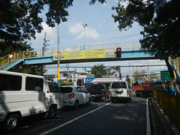 Eine belebte Straße mit Fahrzeugen, eine Brücke mit Geländern und Treppen, Laternen, Verkehrsampeln, Texttafeln, Bäume, Gebäude und ein bewölkter Himmel.
