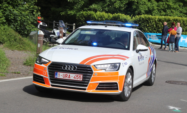 Ein weißes, oranges und blaues Polizeiauto mit sichtbarem Kennzeichen fährt an einem Motorrad auf einer Straße vorbei, mit einer Person im Auto und mehreren Personen in der Nähe; Bäume, Gras, ein Zaun und eine Fahne sind im Hintergrund zu sehen.