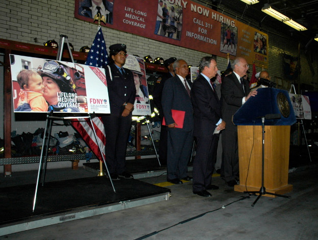 Eine Gruppe von Menschen steht in der Nähe eines Podiums mit Mikrofonen, Schilder auf Ständern, Flaggen und Schilder an der Wand, alles unter hellen Lichtern.