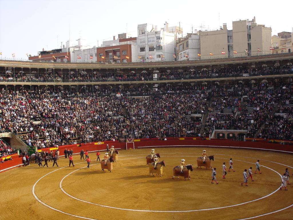 Menschen auf dem Boden gehend, einige reitend auf Pferden und andere sitzend in einem Stadion mit Flaggen, Gebäuden und Himmel im Hintergrund.