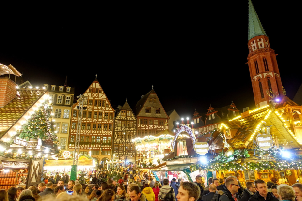 Ein geschäftiger Weihnachtsmarkt in Nürnberg, Deutschland mit Menschen um dekorierte Stände, festliche Lichter, Gebäude mit Fenstern, ein Kirchturm und einen dunklen Himmel.