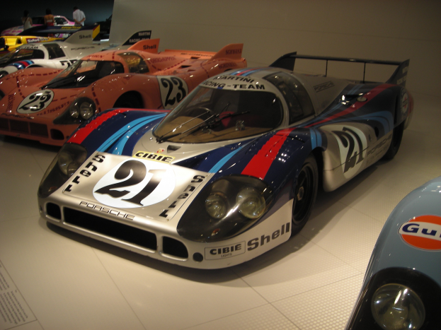 Ein Porsche 917K Le Mans Rennauto in einem Museum ausgestellt, umgeben von anderen Sportwagen und Menschen, mit einer Wand im Hintergrund und einem Blatt Papier mit Text auf der linken Seite.