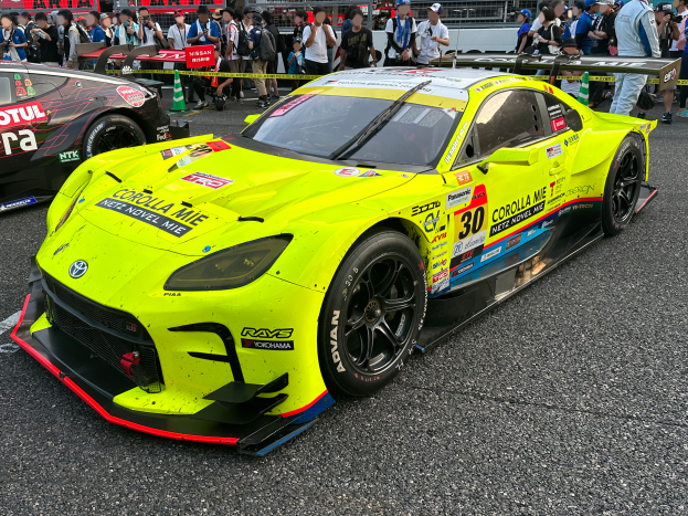 Ein Toyota Supra GT3 Rennwagen auf der Straße, umgeben von einer Menschenmenge, einige machen Fotos, mit Geländern, Schildern und Verkehrskegeln im Hintergrund unter einem klaren blauen Himmel.