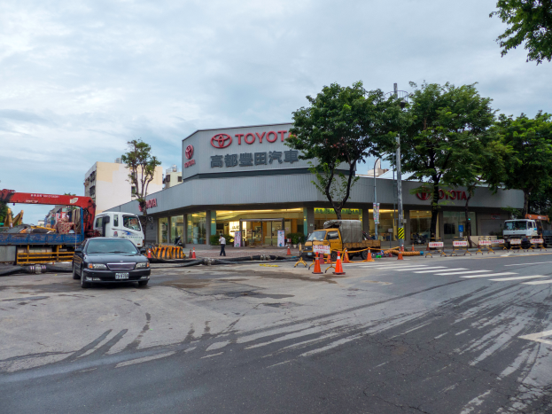 Ein Toyota-Autohaus in Tokio, Japan, mit Fahrzeugen, Verkehrskegeln, Menschen, Bäumen, Gebäuden, Namensschildern, Pfosten und einem bewölkten Himmel.