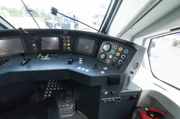 Cockpit eines Zuges mit Feuerlöscher im Vordergrund, das eine Glasfensteransicht von Menschen, Fahrzeugen, Bäumen, Mästäben und anderen Gegenständen außerhalb zeigt.