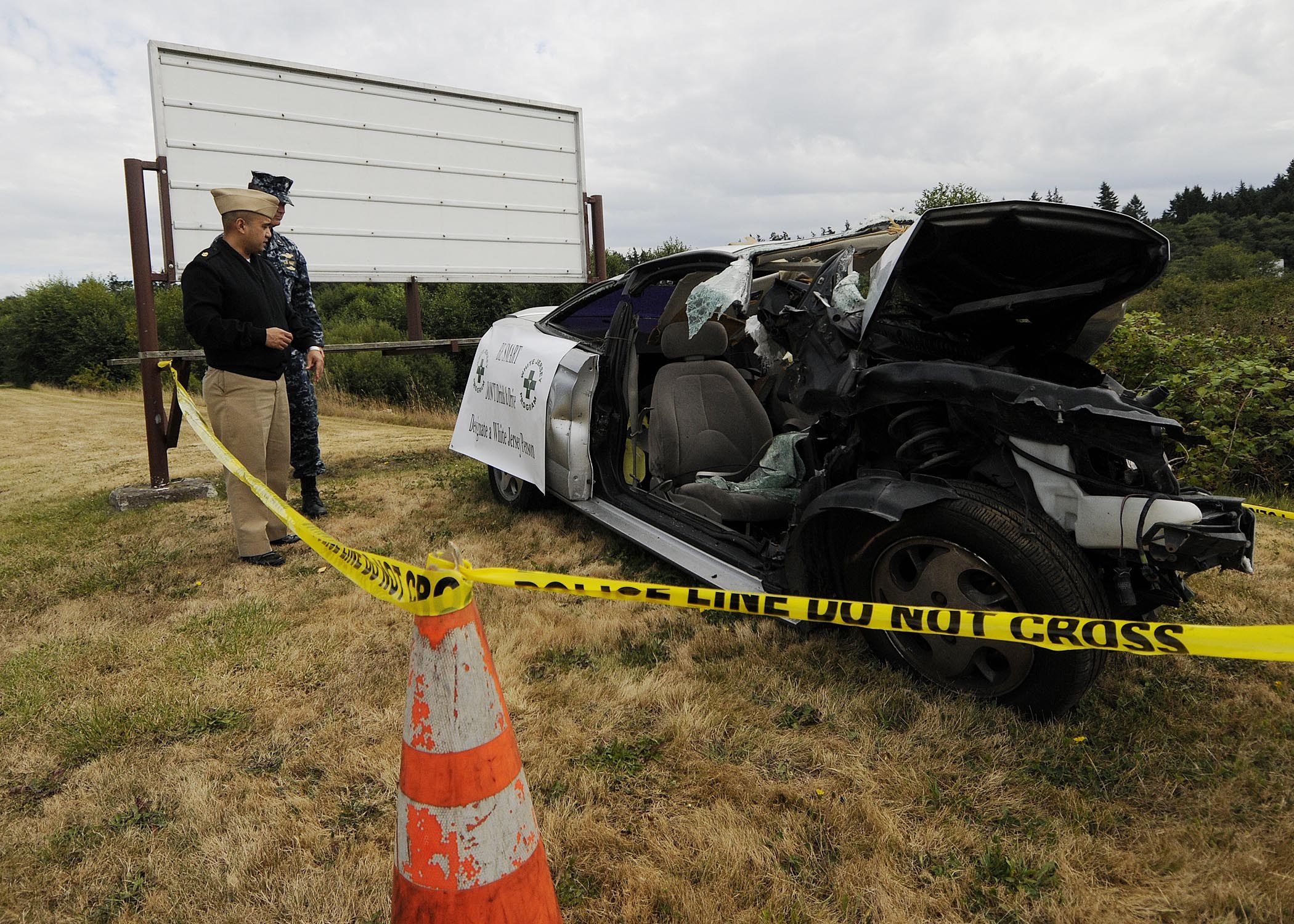 Polizeibeamter in Uniform inspiziert ein schwerbeschädigtes Auto mit eingedellter Motorhaube und zersplitterten Fenstern am Straßenrand, mit einem Verkehrskegel und Band in der Nähe vor dem Hintergrund von Bäumen und einem bewölkten Himmel.