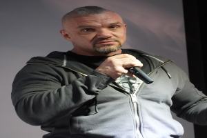 Ein Mann in einer schwarzen Kapuzenjacke und einem schwarzen Anzug mit einem weißen Hemd, der ein Mikrofon hält und selbstbewusst vor einer Wand steht.