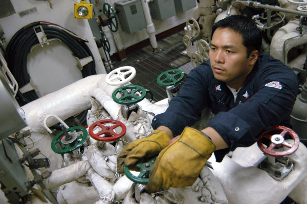 Ein Techniker in einem blauen Hemd und gelben Handschuhen arbeitet an einer Maschine umgeben von Ventilen, Rohren, Drähten und anderem Equipment.