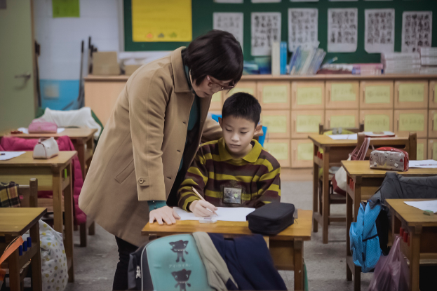 Eine Frau steht neben einem Jungen, der an einem Tisch sitzt und in ein Buch schreibt, in einer Klassenzimmereinrichtung.