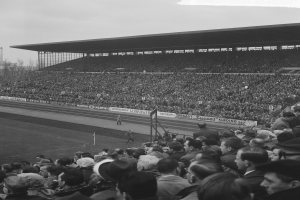 Schwarzes und weißes Foto eines vollen Stadions mit Zuschauern, die ein Fußballspiel verfolgen, einige stehen und andere sitzen auf den Tribünen, mit Bannern, Pfählen, einer Hütte, Bäumen, einem Turm und einem bewölkten Himmel.