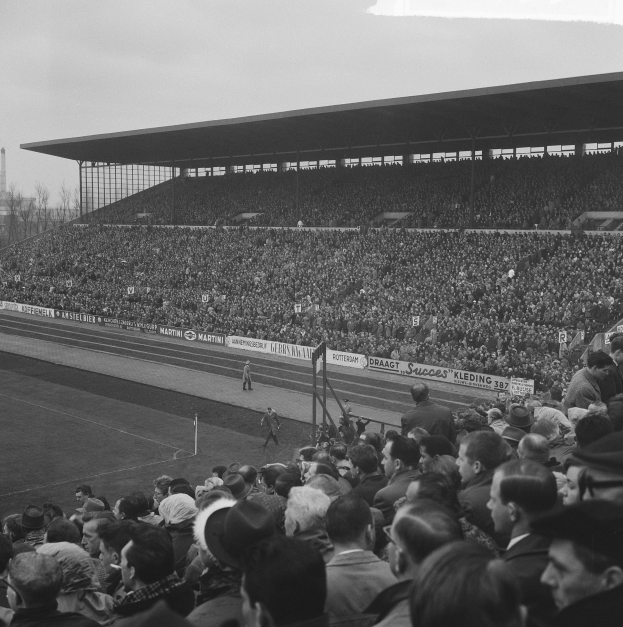 Schwarzes und weißes Foto eines vollen Stadions mit Zuschauern, die ein Fußballspiel verfolgen, einige stehen und andere sitzen auf den Tribünen, mit Bannern, Pfählen, einer Hütte, Bäumen, einem Turm und einem bewölkten Himmel.