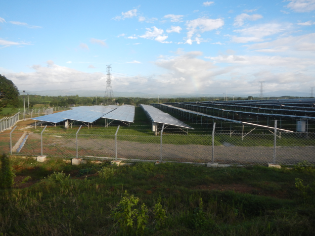 Ein gro├če Solarpark auf einer Wiese umgeben von Pflanzen, einem Zaun, B├Ąumen, ├ťbertragungsmasten und einer bew├Âlktem Himmel.