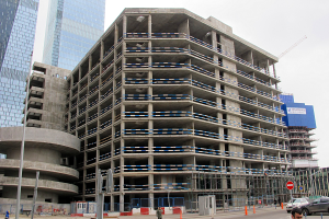 Großes Bauwerk im Bau in einer Stadt, umgeben von Wolkenkratzern, mit Fahrzeugen, Fußgängern, Verkehrsinfrastruktur und Kränen vor einem klaren blauen Himmel.