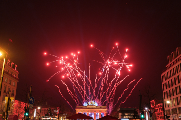 Eine belebte Stadtstraße an einem Silvesterabend in Berlin, mit Gebäuden, Bäumen, Laternenmasten, Verkehrszeichen, Schildern, Zelten, Menschen und einem prächtigen Feuerwerk am Himmel.