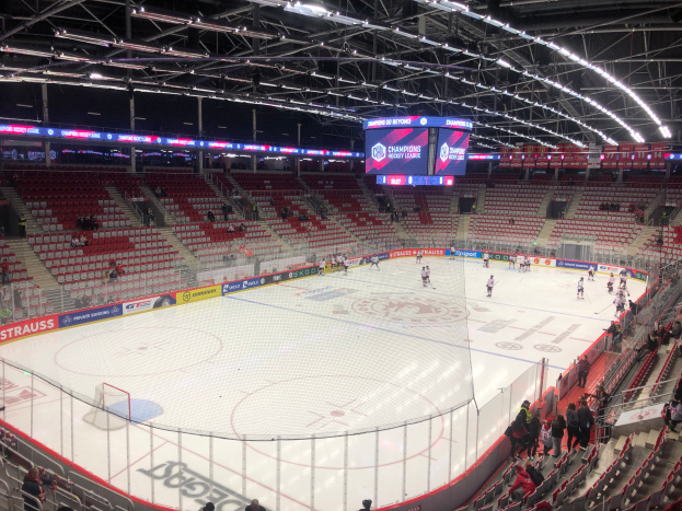 Ein Eishockeyspiel in einer großen Arena mit Zuschauern auf Sitzen und Stehplätzen, einem Zaun um die Eisfläche, einem Display mit der Aufschrift "St. Louis Blues vs Detroit Red Wings", Metallrahmen und einer Decke mit Deckenleuchten.