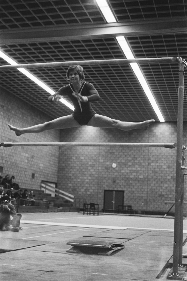 Eine Frau führt eine Gymnastikübung an den Stufenbarren in der Luft aus, mit ausgestreckten Armen und gebeugten Beinen, in einem schwarz-weißen Bild eines Turnhallen mit Zuschauern auf der linken Seite und einer Tür im Hintergrund.