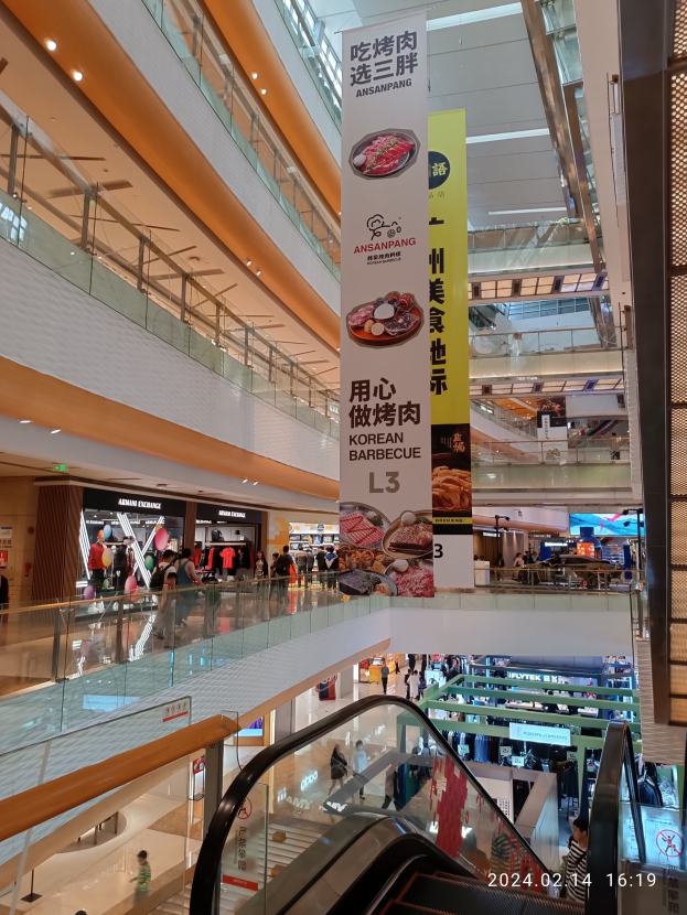 Ein Innenblick in ein Einkaufszentrum mit einer Rolltreppe in der Mitte, Menschen, die herumlaufen, einige mit Handtaschen, und Schilder an der Decke hängen, mit Geschäften im Hintergrund.