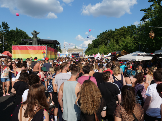 Eine große Menschenmenge marschiert auf einer Straße mit Zelten, Bäumen, Pfählen, Laternen und Gebäuden im Hintergrund bei der Christopher Street Day Parade in Berlin.