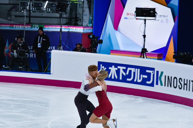 Ein Paar umarmt sich beim Eislaufen auf einer Eisbahn, mit ein paar Menschen im Hintergrund, einige halten Kameras, und eine Tafel mit Schrift darauf, zusammen mit Lichtern und anderen Gegenständen.
