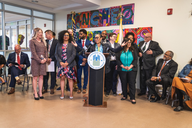 Gruppe von Menschen vor einem Podium mit Mikrofon und einer Tafel mit Text, einige sitzen auf Stühlen und andere stehen, mit Flaggen, einer Tafel, einem Feuerlöscher und Deckenleuchten an der Wand.