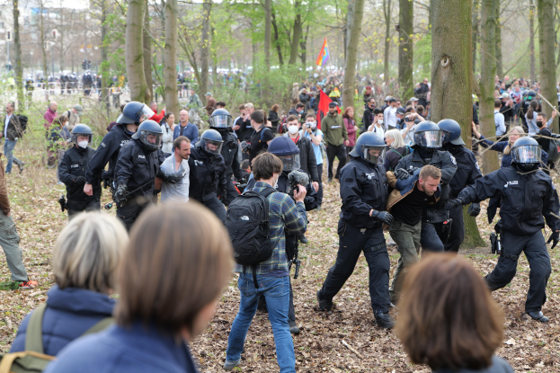 Eine Gruppe von Menschen in Schutzausrüstung steht vor einer Menge von Menschen mit Helmen und Taschen, mit Bäumen, einer Fahne und Gebäuden im Hintergrund und Blättern auf dem Boden.