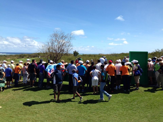 Eine Gruppe von Menschen auf einem grünen Feld bei einem Golfturnier, mit Bäumen, Pflanzen, einer bewölkten Himmel und einem Gewässer im Hintergrund.
