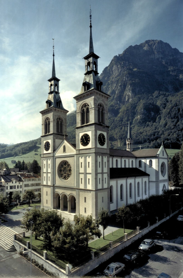 Alte Kirche mit zwei Türmen umgeben von Gebäuden, Grünflächen, Fahrzeugen und Hügeln unter einem bewölkten Himmel.