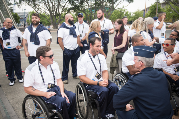 Eine Gruppe von Menschen in Rollstühlen unterhält sich im Freien mit Bäumen, Gebäuden und einem klaren Himmel im Hintergrund, einige tragen Mützen und Schutzbrillen.