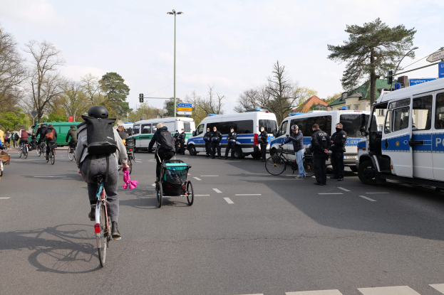 Eine Gruppe von Menschen, die auf Fahrrädern eine Straße entlangfährt, mit Polizeiwagen und anderen Fahrzeugen, einige tragen Helme und haben Taschen oder schieben Kinderwagen, mit Bäumen, Laternenmasten, Verkehrsampeln, Schildern, Gebäuden und einem bewölktem Himmel im Hintergrund.