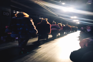 Eine Gruppe von Menschen läuft über den Laufsteg einer Modenschau, während Zuschauer auf beiden Seiten sitzen.