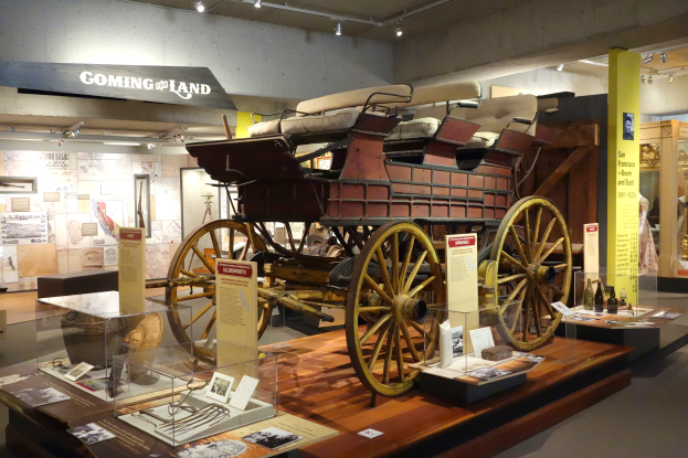 Ein altmodisches Pferdegespann, das in einem Museum ausgestellt ist und von Glasvitrinen mit verschiedenen Gegenständen umgeben ist, mit einer Texttafel an der Wand dahinter, Puppen in Kleidern auf der rechten Seite und Lichter an der Decke.