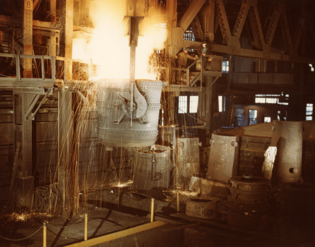 Ein großes Metallobjekt wird in einen Ofen in einer industriellen Umgebung gegossen, umgeben von Arbeitern und beleuchtet vom Feuer des Ofens.