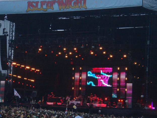 Eine Bühne mit Scheinwerfern, einem Bildschirm und Musikern, die Instrumente spielen, mit einem Banner oben, einer Menge unten und Fahnen mit Stangen und Ballons drum herum.