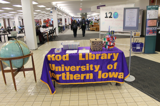 Eine Ausstellung mit einem Tisch, auf dem verschiedene Landesflaggen, eine Kiste, Papiere und ein Banner liegen, eine Kugel links, ein Anschlagbrett rechts mit einigen Leuten in der Nähe und mehreren Tischen und Stühlen links.