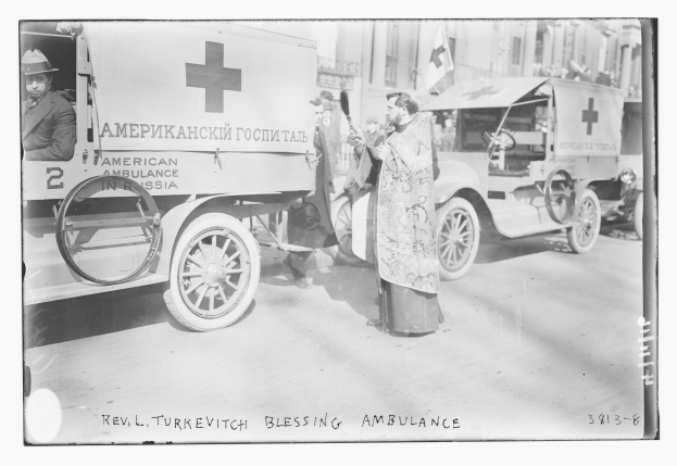 Schwarz-weiß-Foto eines Mannes neben einem frühen 20. Jahrhundert Krankenwagen auf einer Straße mit anderen Fahrzeugen und Menschen, Gebäuden und einer Flagge im Hintergrund und Text unten.