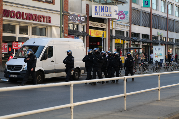 Eine Gruppe von Polizisten in schwarzen Uniformen und Helmen geht √∂ffentlich neben einem weißen Van, mit Fahrr√§dern auf der Seite; Geb√§ude mit Glasfenstern und Laternen sind im Hintergrund zu sehen.