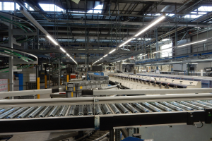 Großer Fabrikinnenraum mit Förderband, helles Oberlicht und mehrere Maschinen und Gegenstände, die auf eine hektische Produktionsumgebung hinweisen.