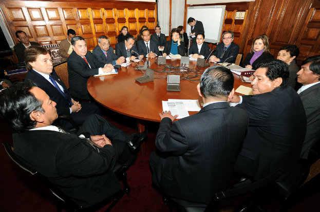 Eine Runde-Tisch-Besprechung mit Delegierten, die diskutieren, Mikrofone, Papiere, Kabel und Dateien auf dem Tisch sowie eine hölzerne Wand im Hintergrund.