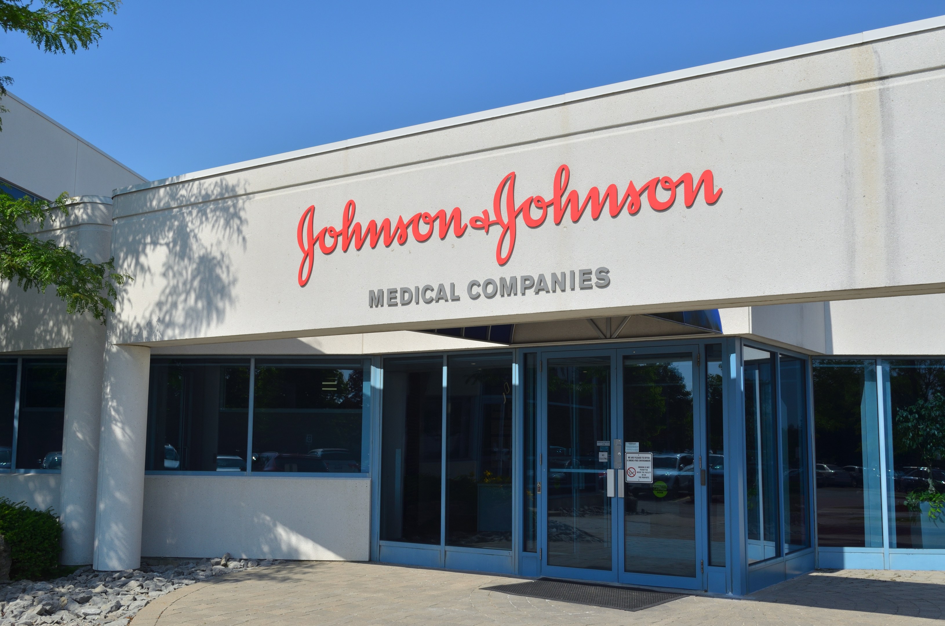 Außenansicht des Johnson & Johnson Medical Companies-Gebäudes mit Glaswänden und -türen, Text an der Wand, Steinen, Pflanzen und einem Baum auf der linken Seite, unter einem klaren blauen Himmel.